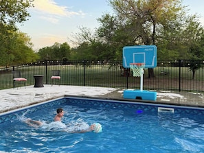 Ein Swimmingpool mit Basketballkorb, umgeben von einem Zaun und Bäumen.