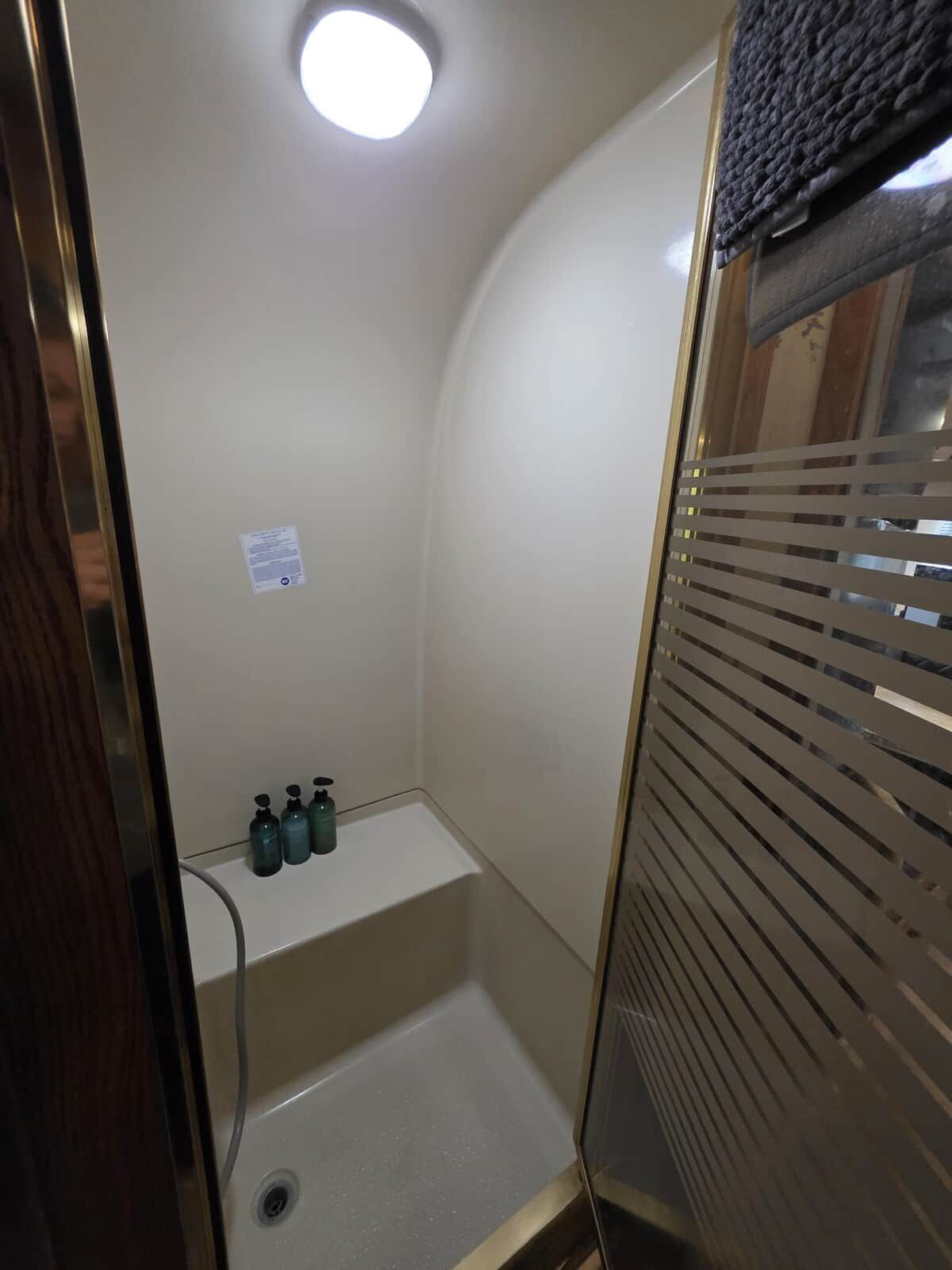 A small, enclosed shower area with a built-in shelf for toiletries and a window with horizontal blinds.