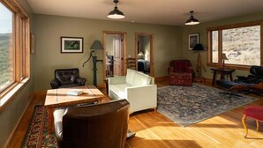 A spacious living room with a wooden floor, a dining table, a sofa, and a large window offering a view of the outdoors.