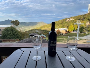A bottle of wine and two glasses on a wooden table with a view of a hilly landscape and buildings in the background.