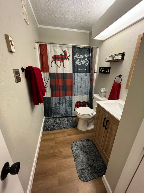 A bathroom with a shower curtain featuring a moose and trees design, a toilet, wooden cabinets, and a small shelf with toiletries.