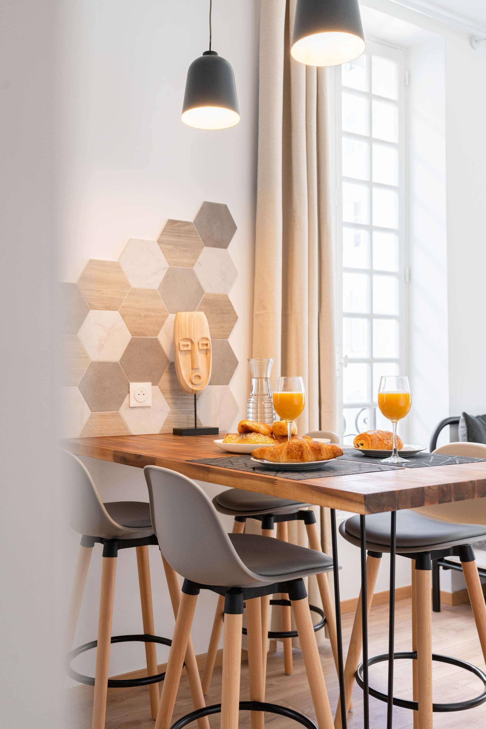 Un espace repas moderne avec une table en bois, des chaises noires et un mur orné d’un motif hexagonal.