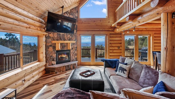 A log cabin interior with a stone fireplace, a flat-screen TV, a sofa with cushions, and large windows offering a view of the outdoors. - Log Cabin near Historic Cripple Creek, Hot Tub and Spectacular Views! (Cripple Creek)