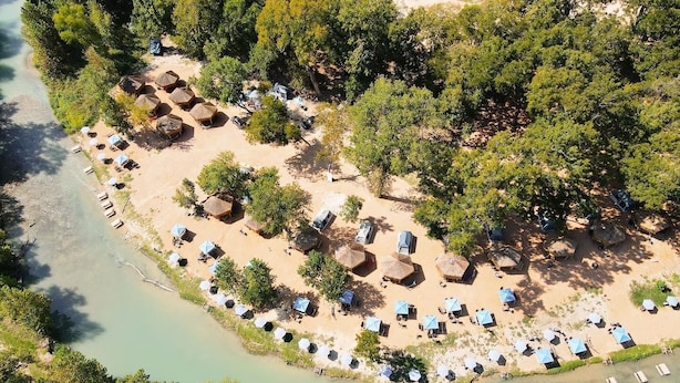 Vista aérea de un complejo turístico en la playa con cabañas de techo de paja y tumbonas dispuestas en filas a lo largo de la orilla.