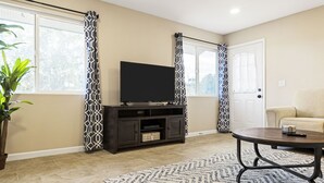 A living room with a flat-screen TV on a dark wooden stand, a beige sofa, a patterned rug, and a potted plant.