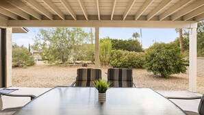 A covered patio with a glass table and two chairs, surrounded by a gravel area and various plants.