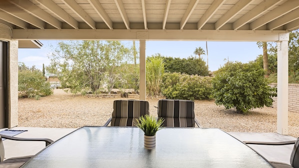 Un patio coperto con un tavolo in vetro e due sedie, immerso in un'area ghiaiata e circondato da varie piante.