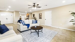 A spacious living room with a ceiling fan, a white sofa with blue and white cushions, a coffee table, and a patterned rug.