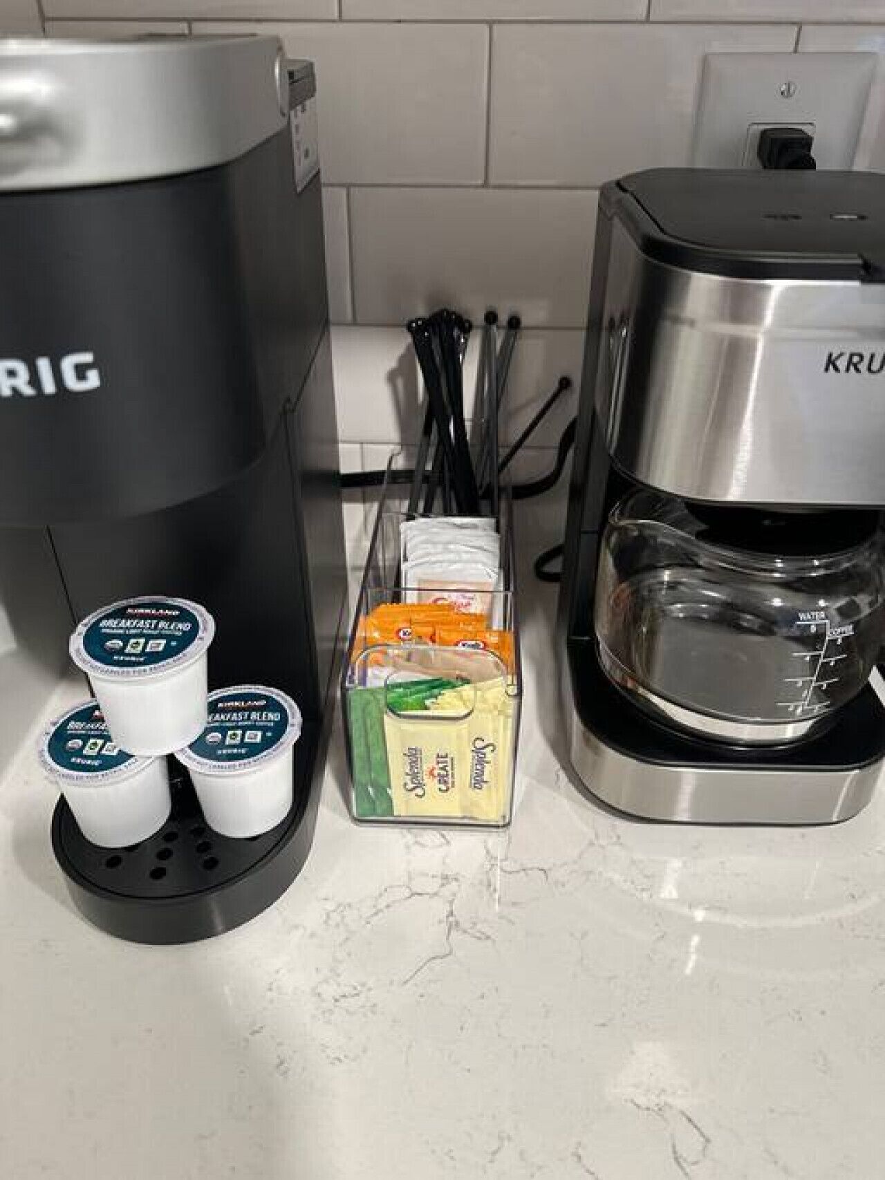 A coffee maker, a Krups coffee machine, and a container with coffee pods and creamer.