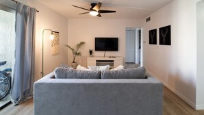 A modern living room with a grey sofa, a ceiling fan, a flat-screen TV, and a wall-mounted TV.