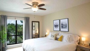 A bedroom with a large bed, a ceiling fan, a nightstand with a lamp, a window with curtains, and a view of greenery outside.