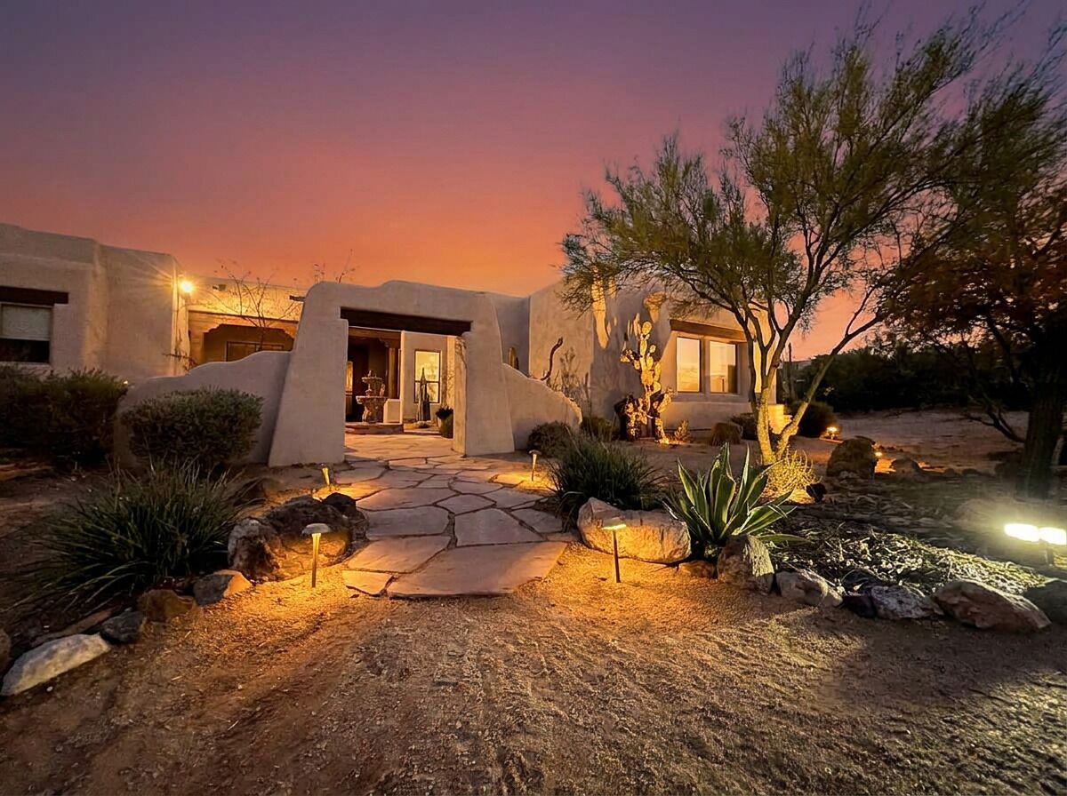 A desert-style house with a stone pathway leading to the entrance, surrounded by native plants and a twilight sky.