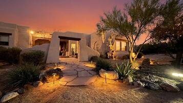 A desert-style house with a stone pathway leading to the entrance, surrounded by native plants and a twilight sky.