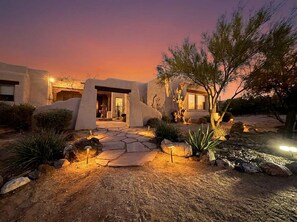 A desert-style house with a stone pathway leading to the entrance, surrounded by native plants and a twilight sky.