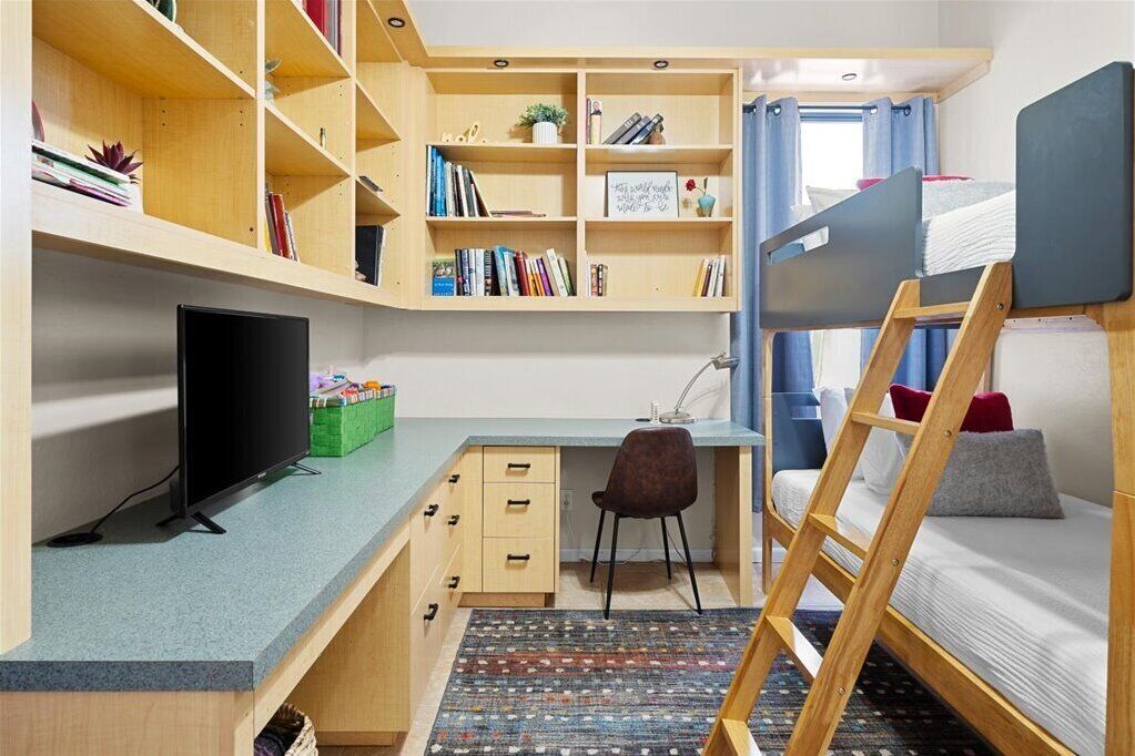 A compact room with a bunk bed, a desk with a computer, a chair, and built-in shelves with books and decorative items.