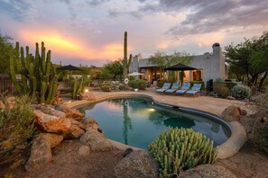 A pool surrounded by rocks and cacti, with a modern house in the background.