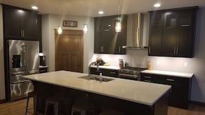 A modern kitchen with dark cabinetry, stainless steel appliances, a central island with stools, and pendant lighting.