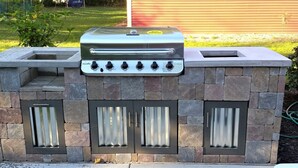 Ein Stein-Barbecue-Grill mit vier Brennern und einem Schornstein, vor einem Haus mit Rasengarten.
