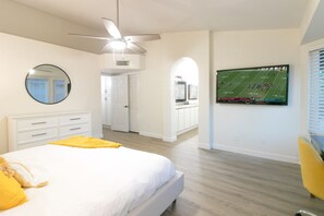 A bedroom with a bed, a ceiling fan, a TV mounted on the wall, and a hallway leading to a bathroom.