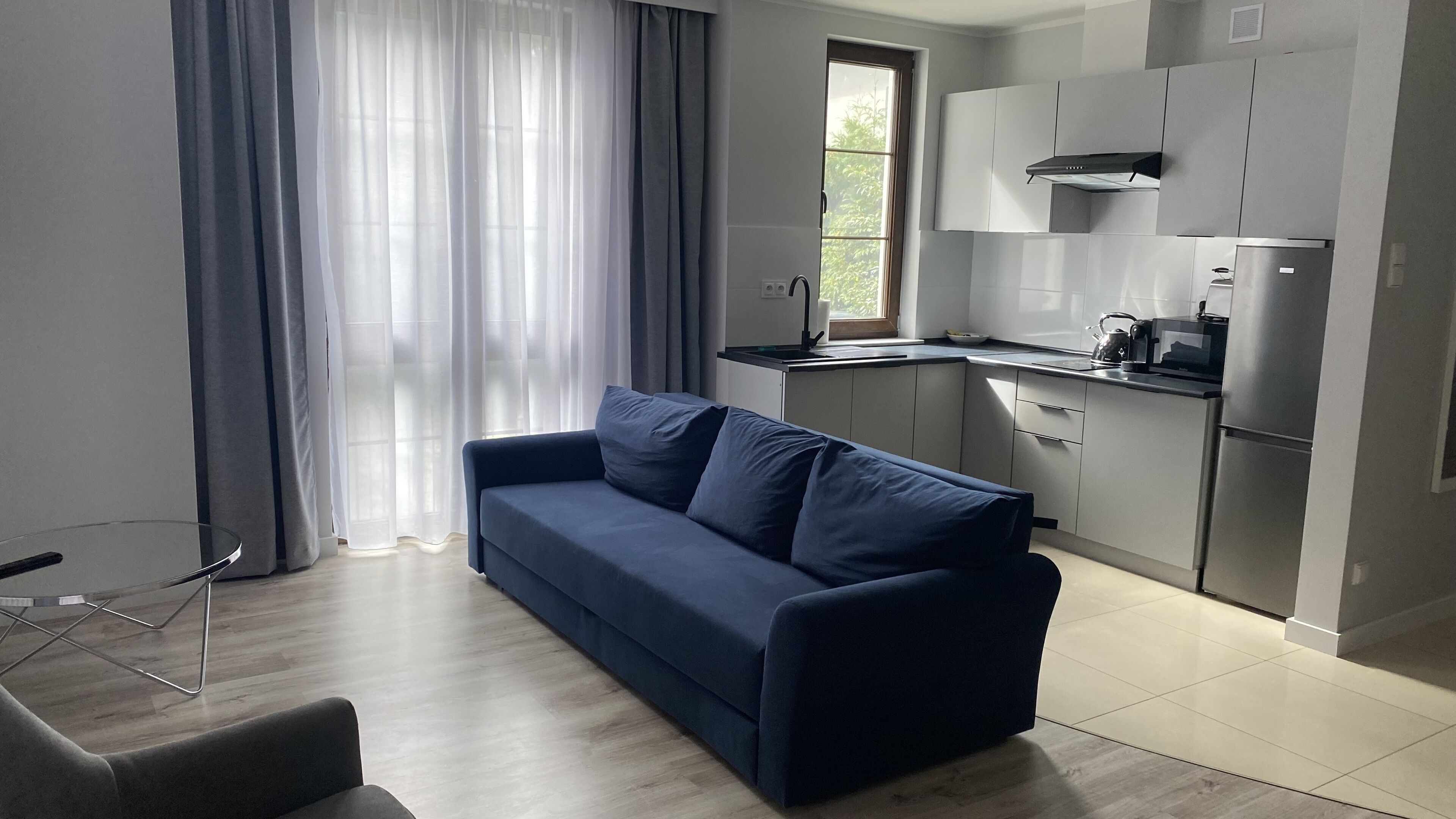 Un salon moderne avec un canapé bleu foncé et une table basse en verre. Attenant au salon se trouve une cuisine avec des armoires blanches et un réfrigérateur en acier inoxydable.