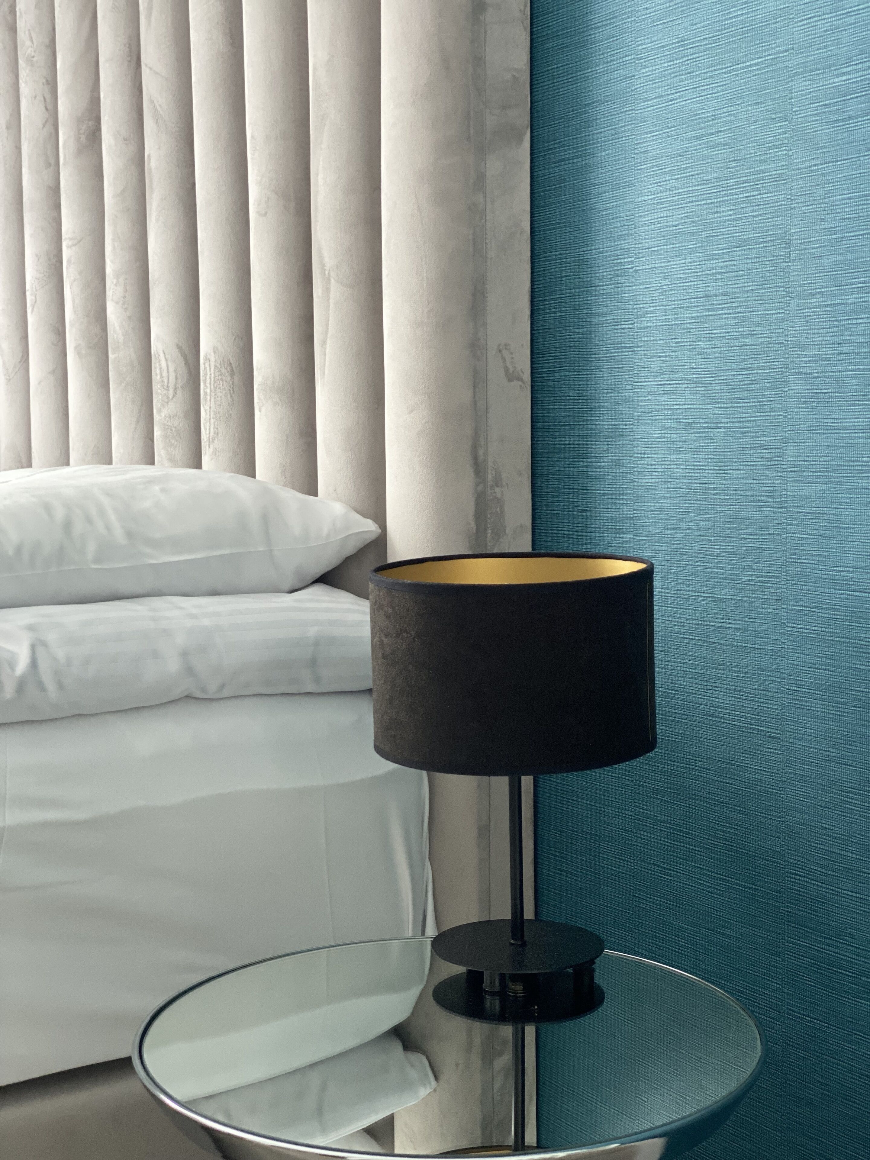 A modern bedroom with a bed, a round glass side table, a black lamp, and a blue textured wall.