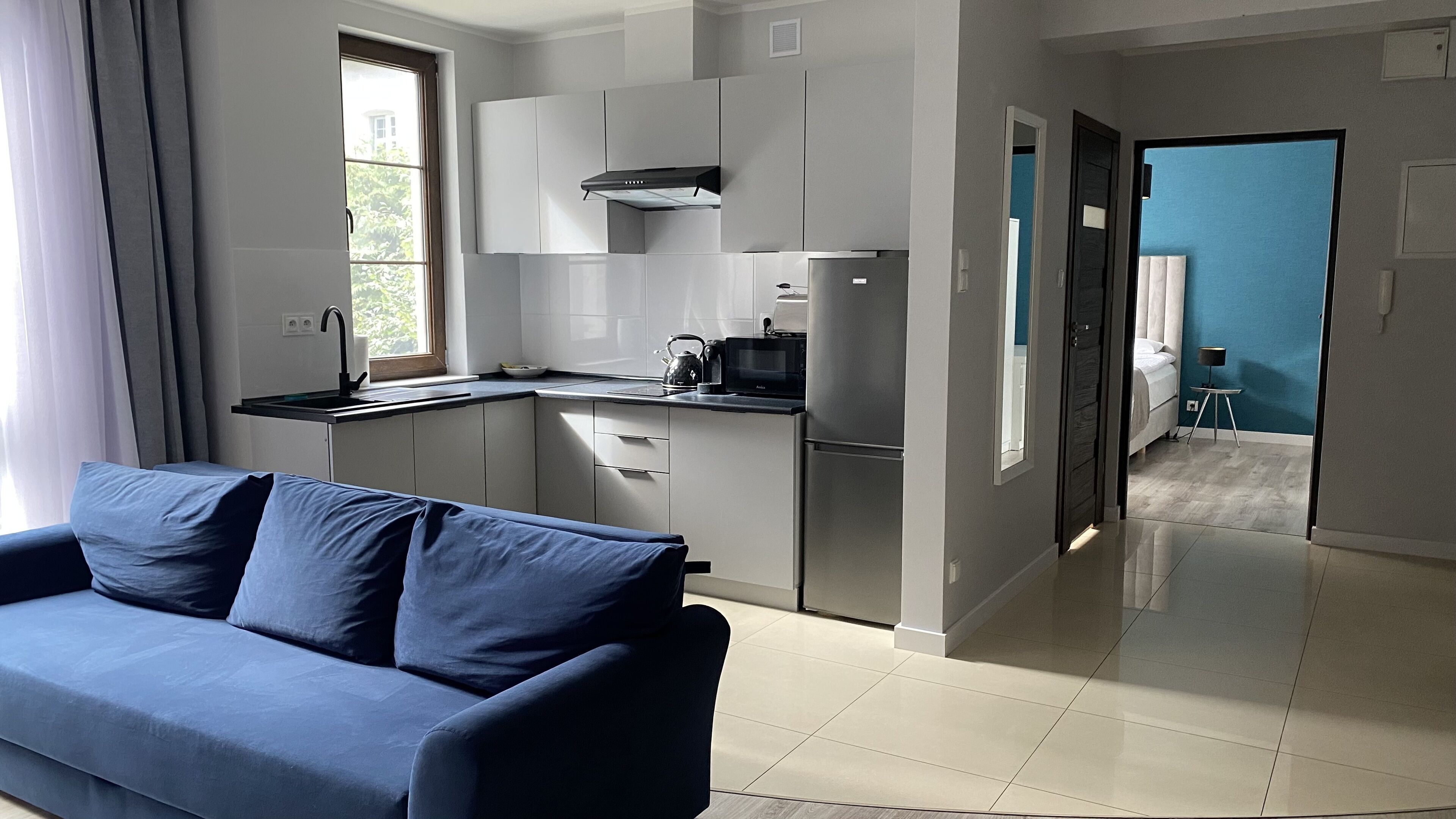 A modern kitchen with grey cabinets, a black microwave, and a kettle. There is a blue sofa in the living area.