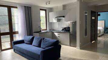 Cozinha moderna com forno embutido, micro-ondas e pia. Há um sofá azul na sala de estar e uma cama no quarto vizinho.