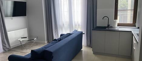 Un espacio habitable moderno con sofá, una mesita, televisor, zona de cocina con fregadero y ventana con cortinas.