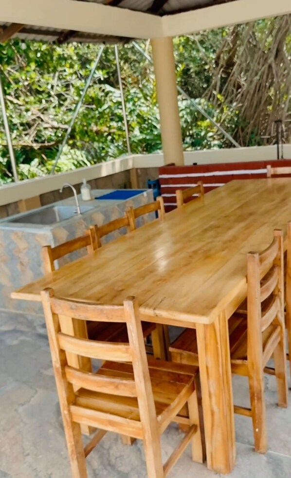 A wooden dining table with chairs, a bench, and a view of greenery through a covered area.