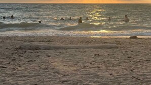 Una playa con un atardecer sobre el océano, una orilla arenosa y gente nadando.