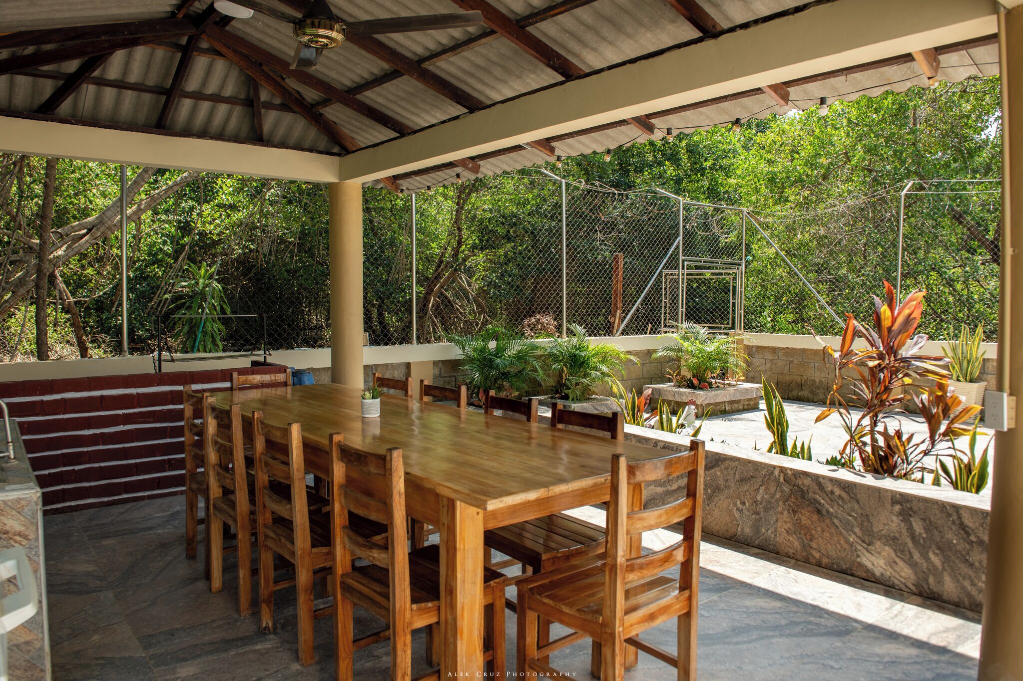 An outdoor dining area with a wooden table and chairs, surrounded by potted plants and a stone wall.