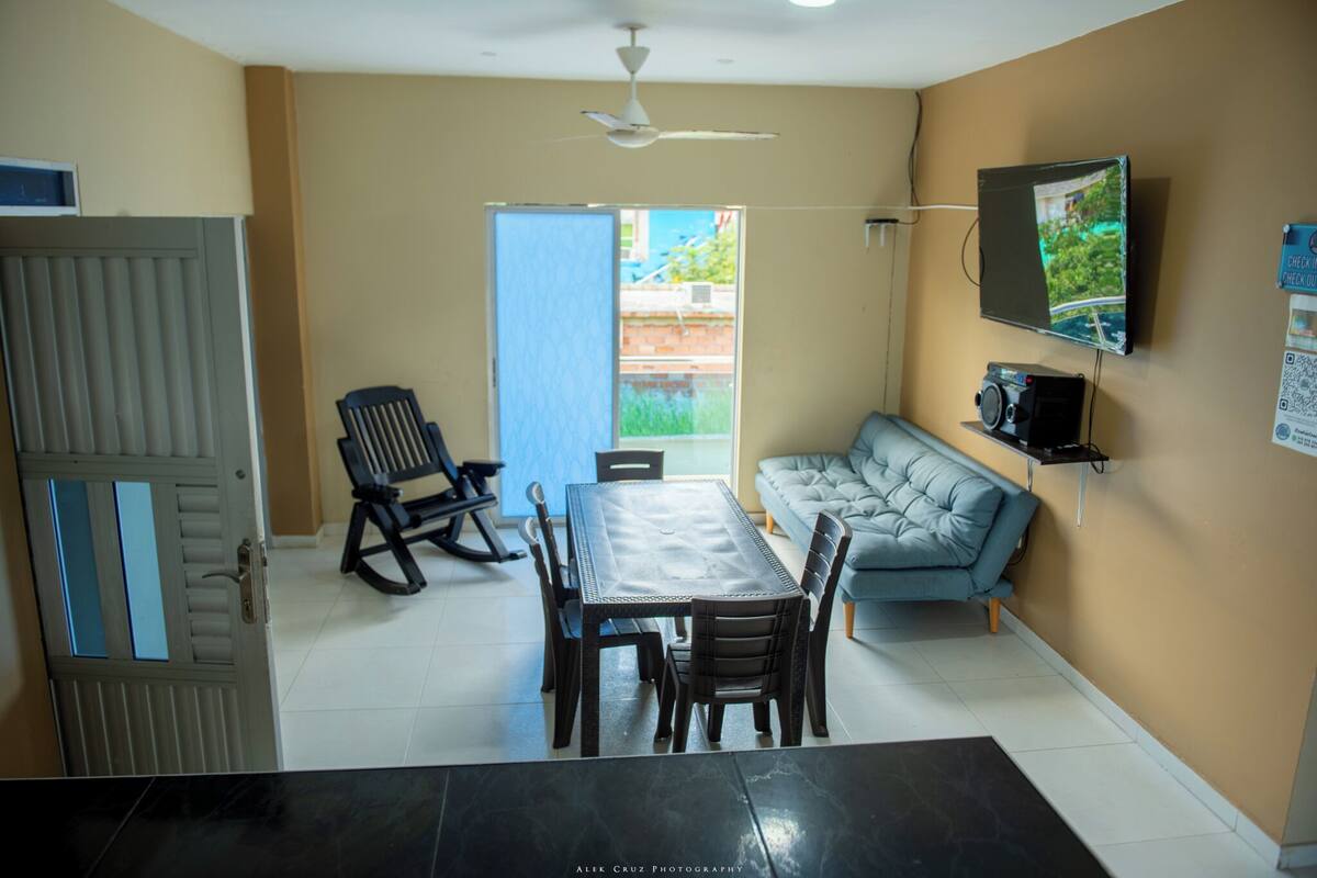 Un comedor con mesa y sillas, un sofá, un ventilador de techo y un televisor montado en la pared.