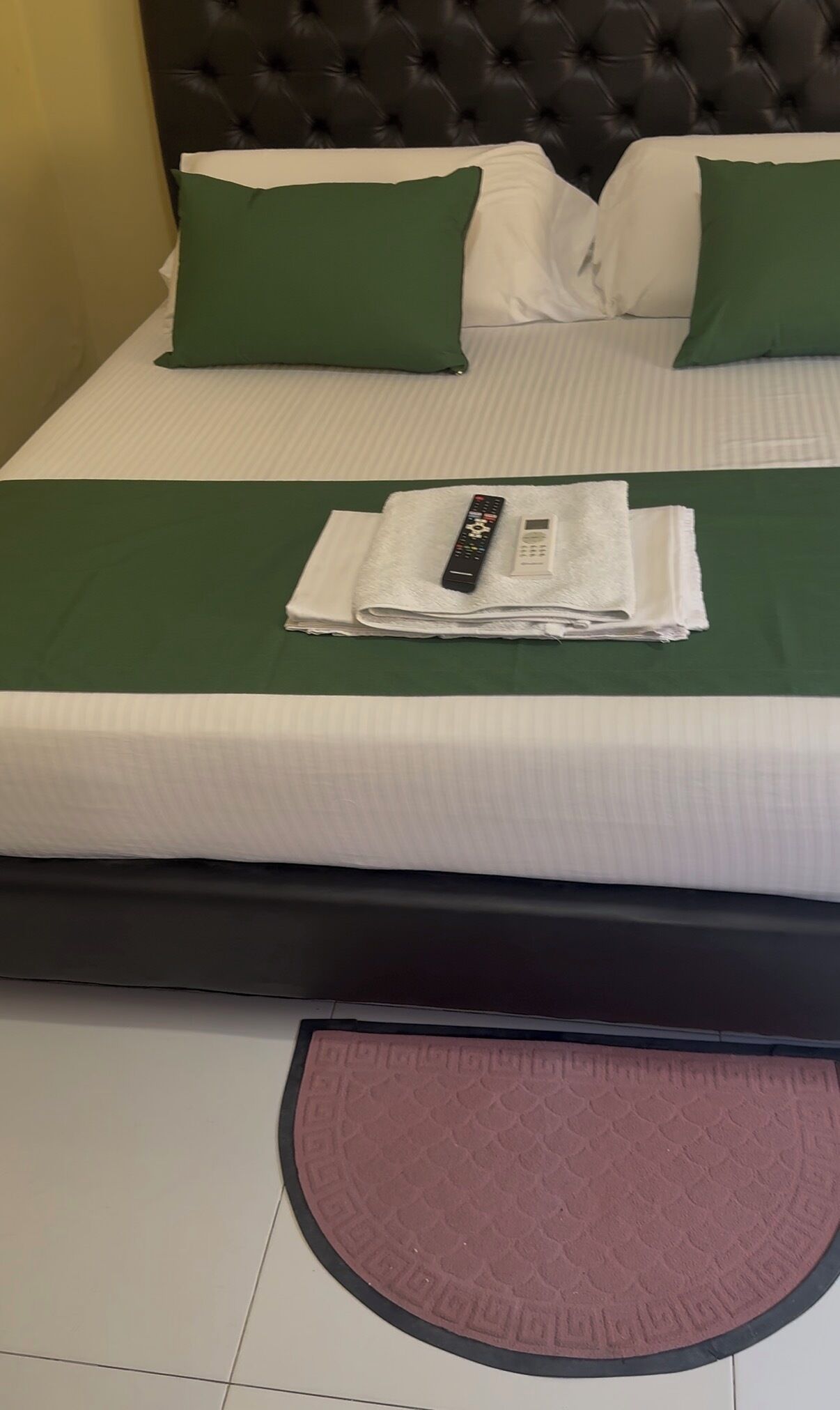 A neatly made bed with green pillows, a folded white towel, and a remote control on the bed.