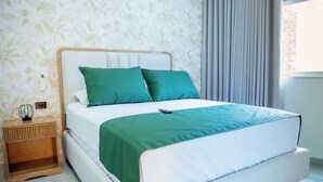 Una cama bien tendida con cabecera blanca y ropa de cama verde, una mesita de noche con lámpara y una ventana con cortinas.
