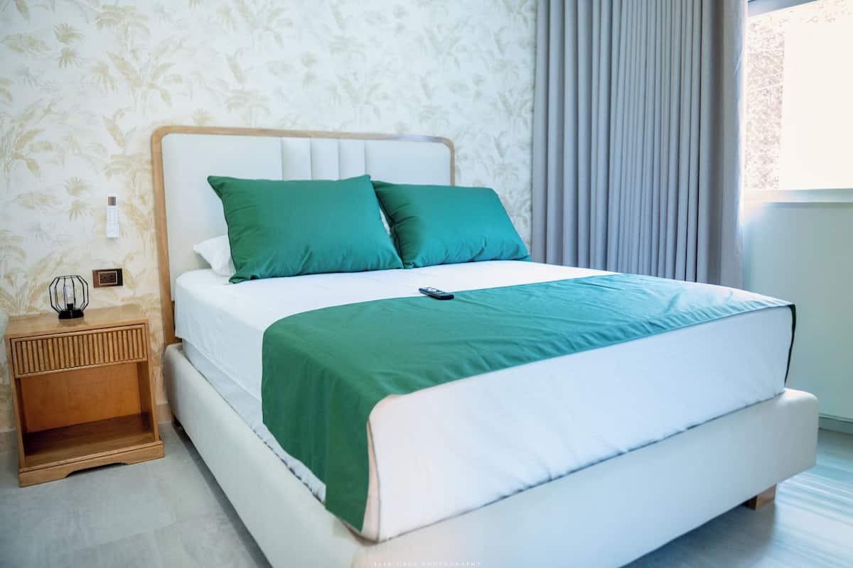 Una cama bien tendida con cabecera blanca y ropa de cama verde, una mesita de noche con lámpara y una ventana con cortinas.