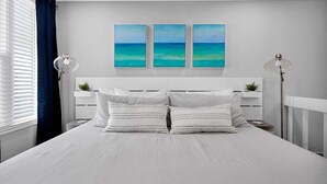 A modern bedroom with a large bed, white bedding, and three wall-mounted paintings depicting a blue ocean. - Beachside! Jacksonville Beach Condo-Close to Mayo Clinic (Jacksonville Beach)