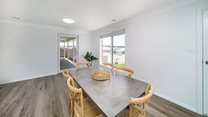 A spacious dining area with a long table and wooden chairs, large windows offering a view, and a potted plant on the table. - Modern Escape in Quiet Neighbourhood (Gobbagombalin)