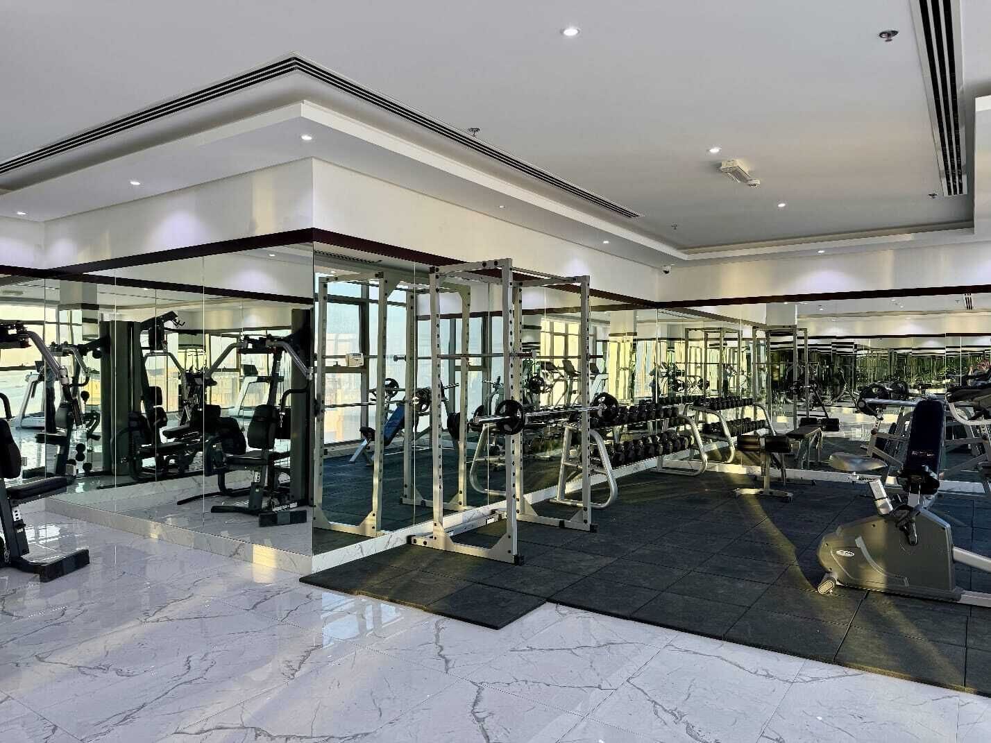 A modern gym with various exercise equipment, large windows, and a polished marble floor.