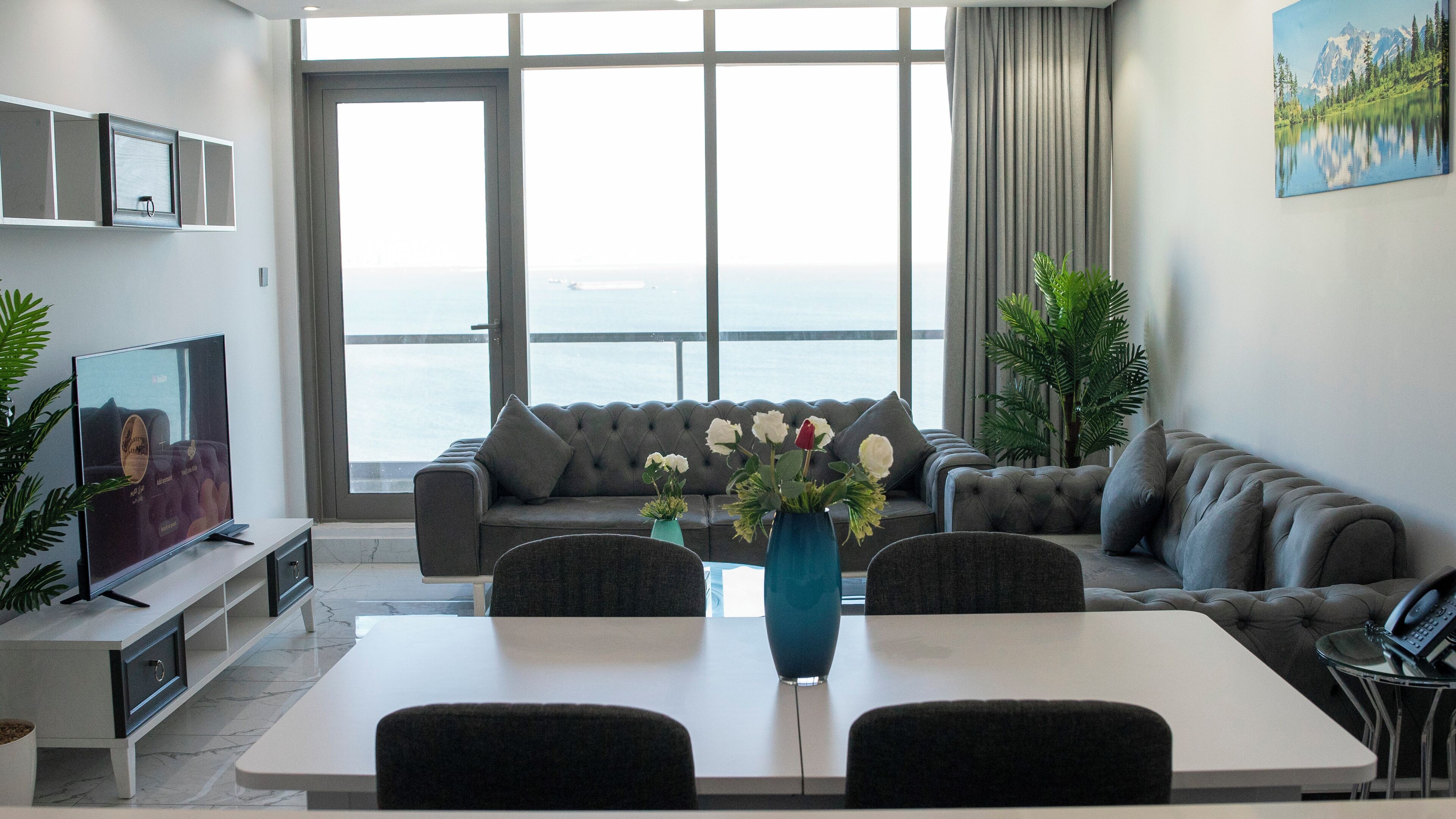 Un salon moderne avec une grande fenêtre, des canapés gris, une table à manger et un téléviseur posé sur un meuble.