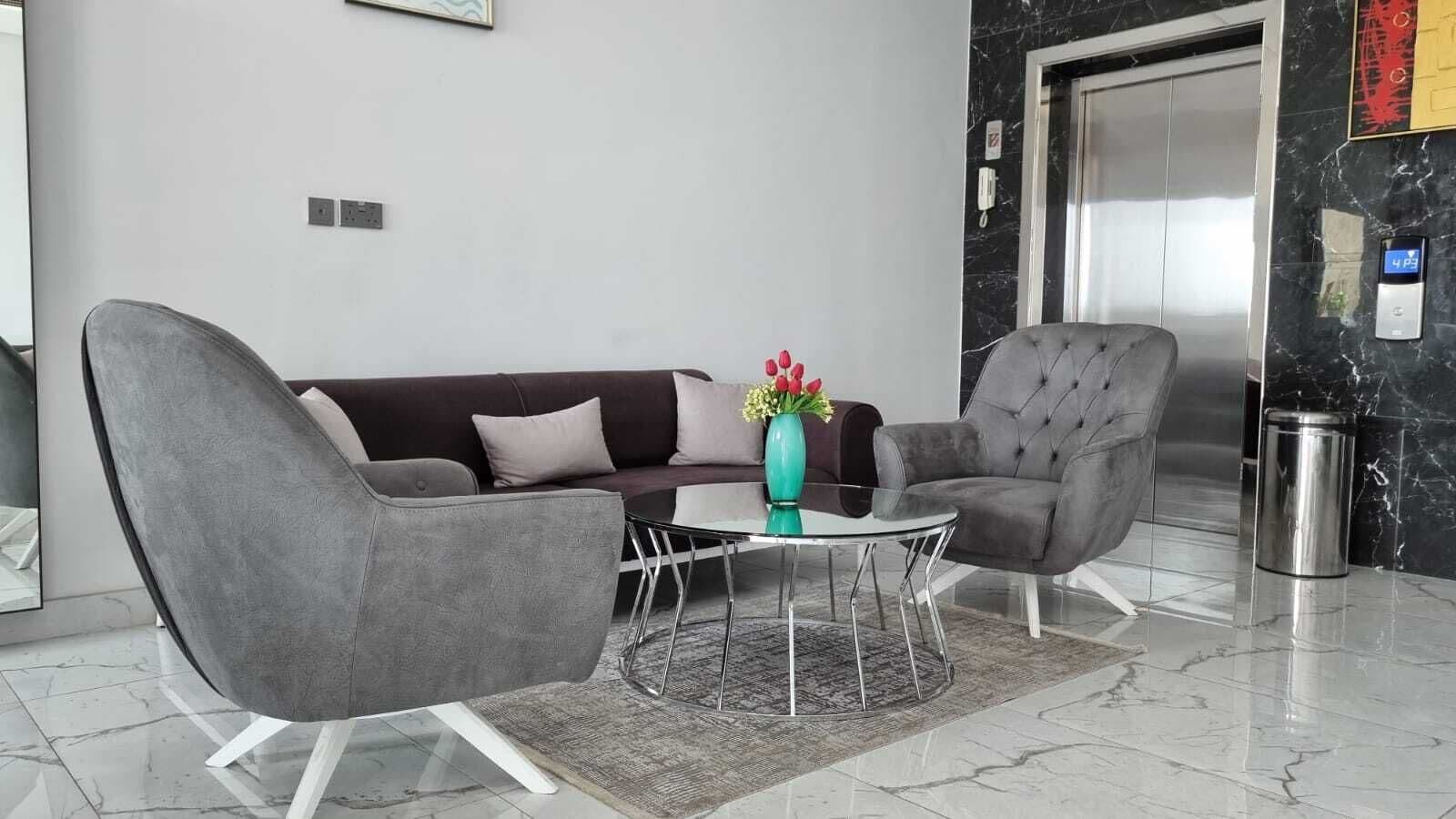 A modern lobby with a grey sofa, a grey armchair, a glass coffee table, and a vase with flowers.