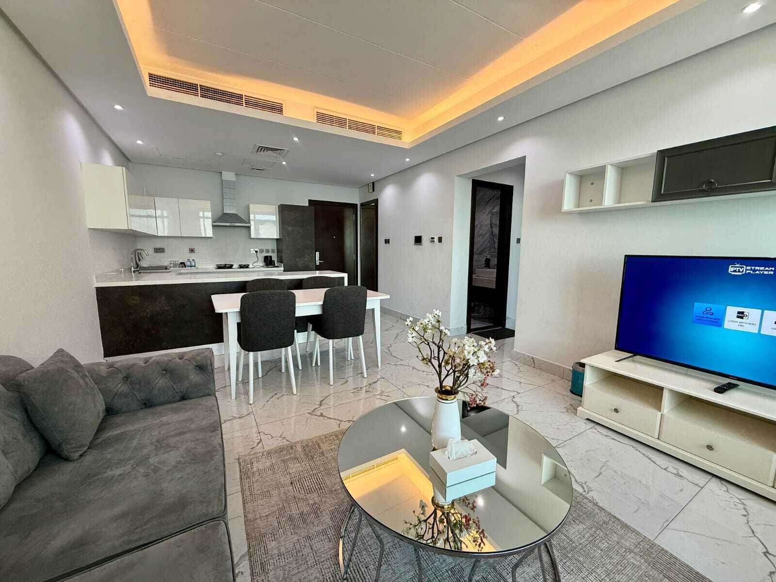 A modern living room with a sofa, a coffee table, and a TV. There is a kitchen area in the background with a dining table and chairs.