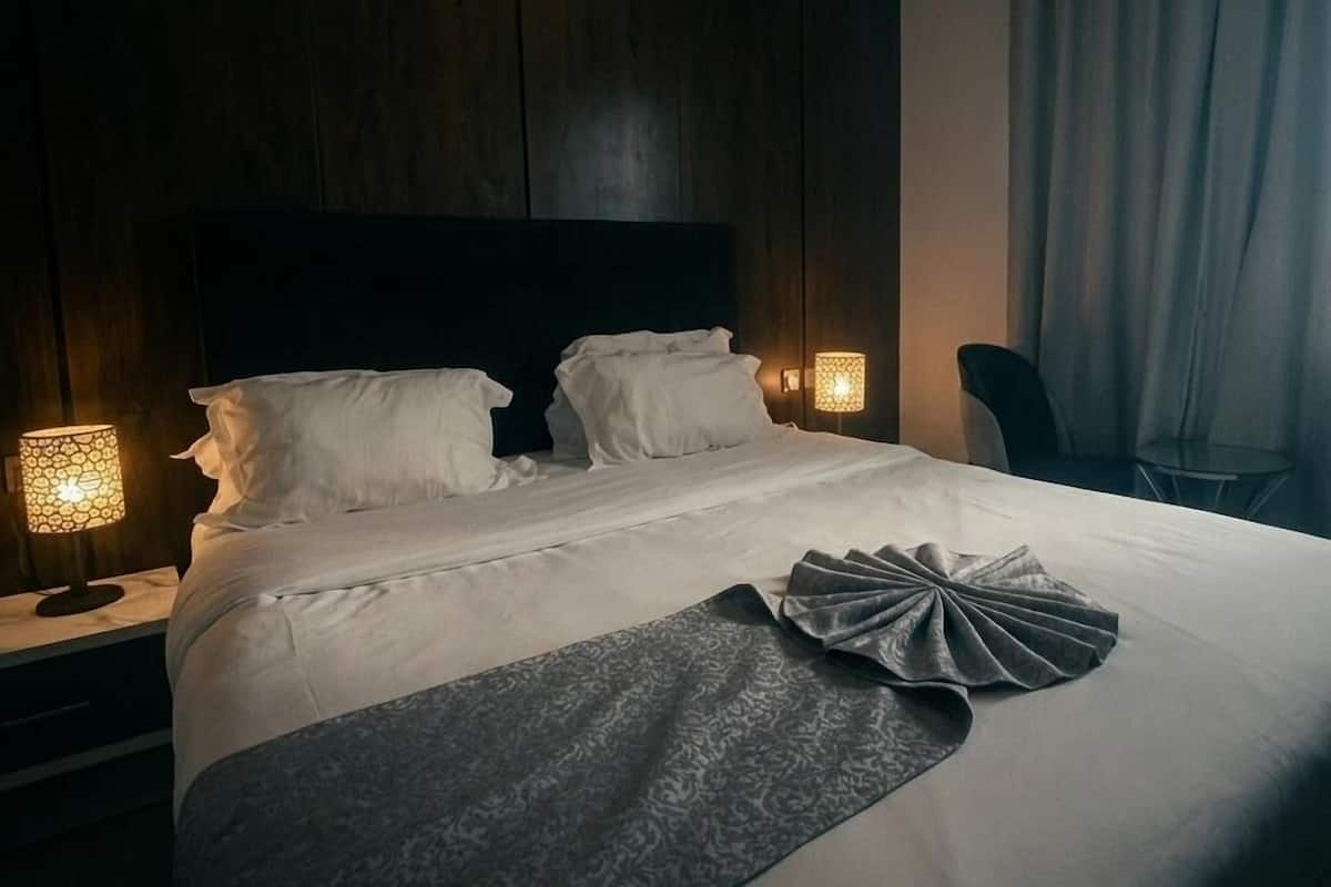 A modern hotel room with a large bed, two bedside lamps, a dark wooden headboard, and a grey throw blanket.