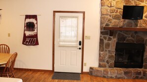 A dining area with a wooden table and chairs, a stone fireplace with a TV above it, and a door with a window. - Winetrail Cabin - SIU, Giant City SP, Shawnee Forest, & Cedar Lake minutes away! (Makanda)