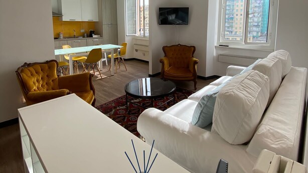 Een moderne woonkamer met een witte bank, gele fauteuils, een glazen salontafel en een aan de muur bevestigde televisie.