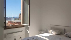 Een slaapkamer met een bed, nachtkastje, raam met een open luik en uitzicht op gebouwen.
