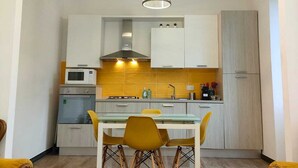 Een kleine keuken met witte kastjes, een magnetron en een gasfornuis. Op de voorgrond staat een eettafel met gele stoelen en een tafel met een glazen blad.