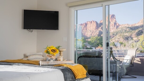 A bedroom with a bed, a flat-screen TV, a vase with sunflowers, and a view of a mountainous landscape through a sliding glass door. - So Zion Painted Desert-4Bed, 3Bath, Hot Tub, Deck (Hildale)
