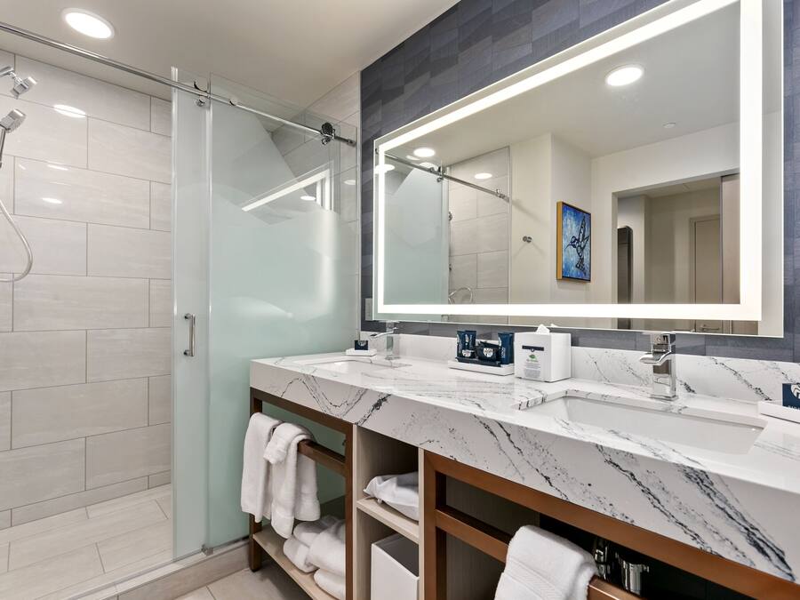 Un baño moderno con ducha envuelta en vidrio, lavamanos dobles y encimeras de mármol, y un espejo grande.
