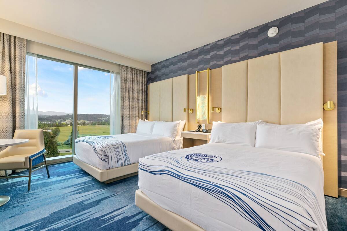 A hotel room with two beds, a large window with a view, a round table, and a modern design.