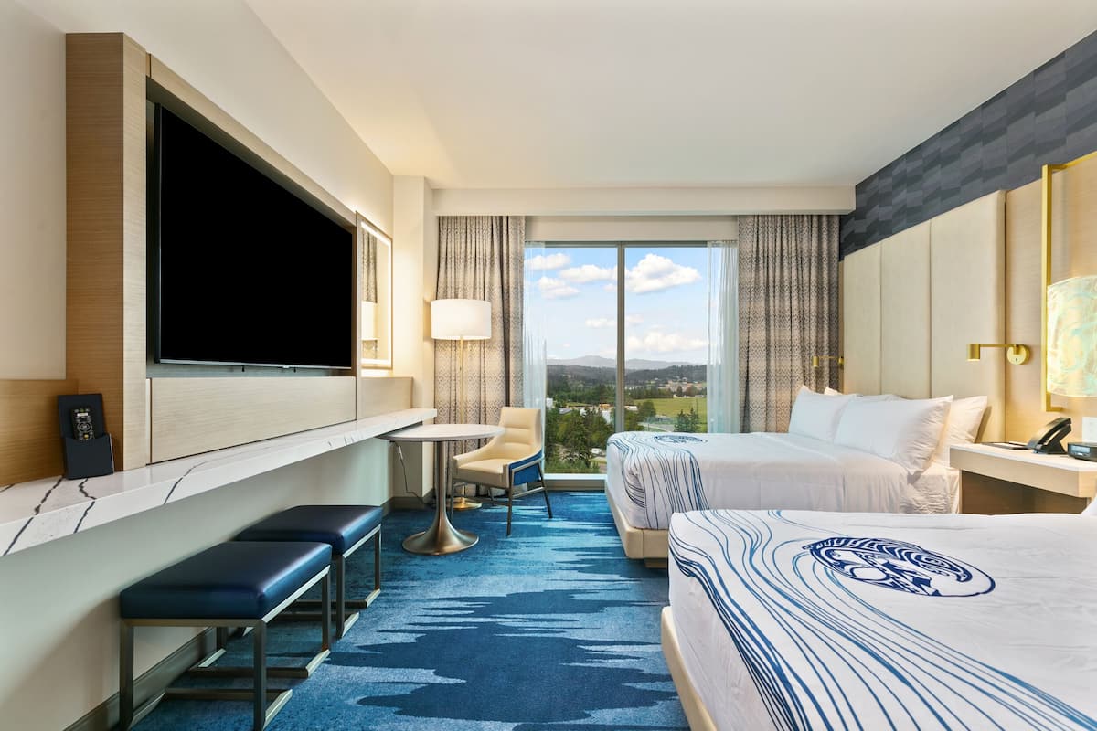 A hotel room with a large TV, a desk with a chair, two beds, and a view of the city.